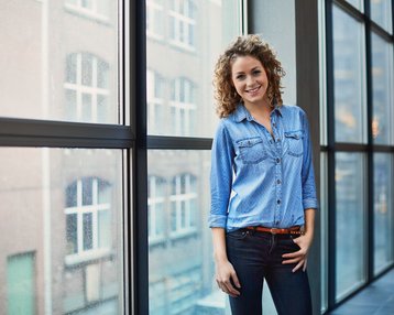 Eine lächelnde Frau mit lockigem Haar steht an einem großen Fenster und trägt ein blaues Hemd und Jeans. Im Hintergrund sind Gebäude sichtbar.