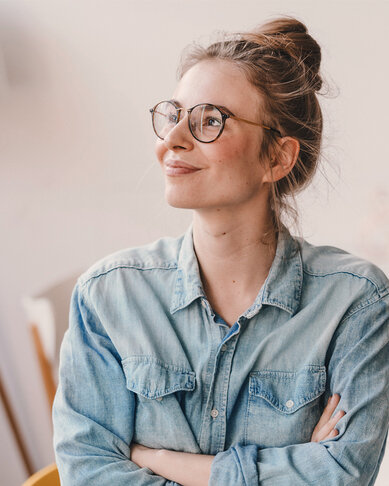 Eine lächelnde Frau mit Brille sitzt in einem hellen Raum, trägt ein Jeanshemd und hat die Arme verschränkt. Sie schaut nachdenklich und wirkt zufrieden.