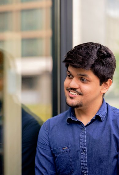 Ein junger Mann mit dunklem Haar sitzt lächelnd vor einem Fenster, das die Umgebung reflektiert. Er trägt ein blaues Hemd und wirkt nachdenklich.
