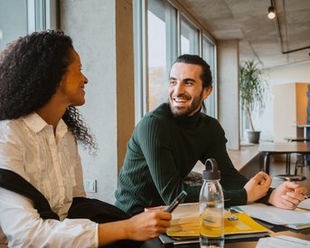 Zwei Personen sitzen an einem Tisch, lächeln sich an und unterhalten sich. Auf dem Tisch liegen Notizblätter und eine Wasserflasche.