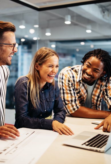 Vier Personen lachen und schauen gemeinsam auf einen Laptop in einem modernen Büro. Sie scheinen an einem Projekt zu arbeiten und genießen die Zusammenarbeit.