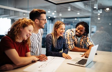 Vier Personen lachen und schauen gemeinsam auf einen Laptop in einem modernen Büro. Sie scheinen an einem Projekt zu arbeiten und genießen die Zusammenarbeit.