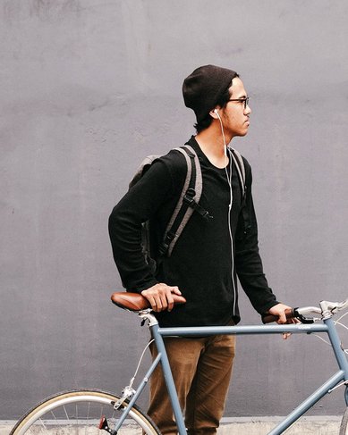Ein junger Mann steht neben einem Fahrrad vor einer grauen Wand. Er trägt einen schwarzen Pullover, eine Mütze und hat einen Rucksack auf dem Rücken.