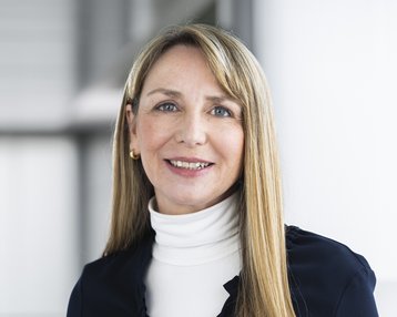 Eine Frau mit langen, glatten Haaren trägt ein dunkelblaues Oberteil und einen weißen Rollkragenpullover. Sie lächelt freundlich.