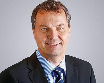 Ein Mann mit kurzen, braunen Haaren und einem freundlichen Lächeln trägt einen schwarzen Anzug und ein gestreiftes Hemd. Der Hintergrund ist neutral und hell.