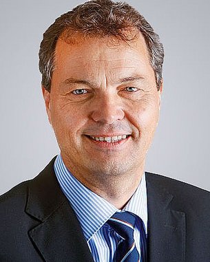 Ein Mann mit kurzen, braunen Haaren und einem freundlichen Lächeln trägt einen schwarzen Anzug und ein gestreiftes Hemd. Der Hintergrund ist neutral und hell.