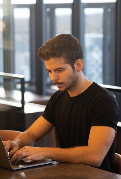Ein junger Mann sitzt an einem Tisch und arbeitet konzentriert an einem Laptop in einem modernen, hellen Raum mit großen Fenstern.