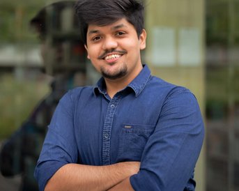 Ein junger Mann mit dunklen Haaren steht lächelnd vor einer Glaswand. Er trägt ein blaues Hemd und hat die Arme vor der Brust verschränkt. Im Hintergrund sind unscharfe Bäume und Gebäude zu sehen.