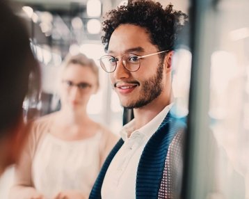 Ein junger Mann mit lockigem Haar und Brille lächelt in die Kamera, während eine Frau im Hintergrund steht und ebenfalls lächelt. Sie befinden sich in einem modernen, hellen Raum.