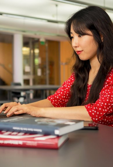 Eine Frau mit langen, dunklen Haaren sitzt an einem Tisch in einem modernen Raum und arbeitet konzentriert an einem Laptop. Auf dem Tisch liegen einige Bücher.