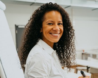 Eine lächelnde Frau mit lockigem Haar steht neben einem Flipchart und hält einen Marker in der Hand. Sie trägt ein weißes Hemd und wirkt freundlich.