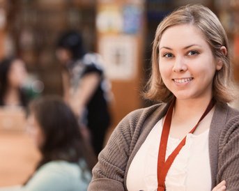 Eine lächelnde Frau mit blonden Haaren steht in einer Bibliothek. Im Hintergrund sind andere Personen zu sehen, die an Computern arbeiten.