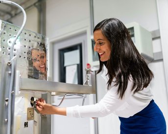 Eine Frau bedient eine Maschine in einem modernen Labor, während ein Kind neugierig durch ein Fenster schaut.