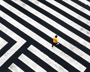 Eine Person in gelber Kleidung geht über ein geometrisches Zebrastreifenmuster aus schwarzen und weißen Linien.