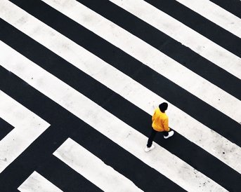 Eine Person in gelber Kleidung geht über ein geometrisches Zebrastreifenmuster aus schwarzen und weißen Linien.