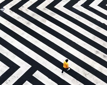 Eine Person in gelber Kleidung geht über ein geometrisches Zebrastreifenmuster aus schwarzen und weißen Linien.