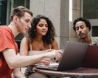 Drei Personen sitzen an einem Tisch und schauen auf einen Laptop. Sie diskutieren und zeigen auf den Bildschirm, während sie konzentriert arbeiten.