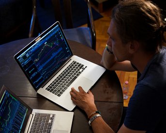Ein Mann sitzt an einem Tisch mit zwei Laptops, die Finanzdiagramme anzeigen. Er schaut konzentriert auf die Bildschirme.