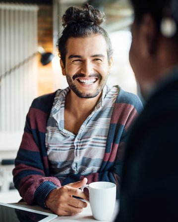 Ein lächelnder Mann sitzt an einem Tisch, hält eine Tasse und spricht mit einer anderen Person. Im Hintergrund sind moderne Möbel und eine helle, einladende Atmosphäre zu sehen.