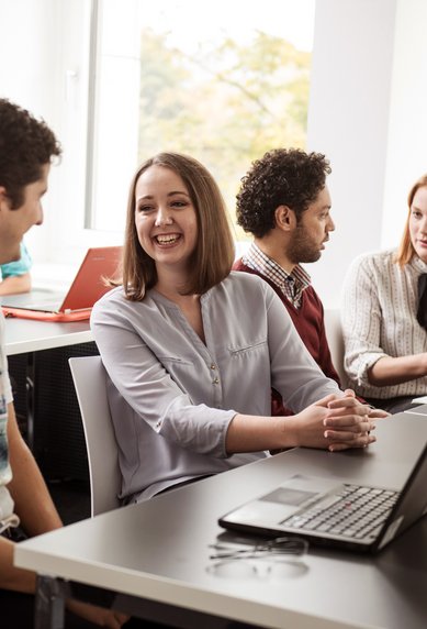 Eine Gruppe von Studierenden sitzt an Tischen, unterhält sich und arbeitet an Laptops in einem modernen Klassenzimmer mit großen Fenstern.