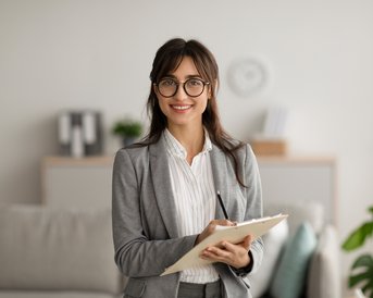 Eine lächelnde Frau in einem grauen Anzug hält ein Notizbuch in einem modernen Büro mit Pflanzen und einem Sofa im Hintergrund.
